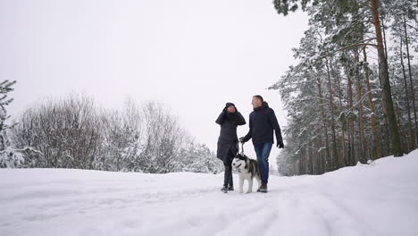 Waist-up-portrait-of-happy-modern-couple-playing-with-cute-Husky-puppy-outdoors-in-winter,-focus-on-Asian-man-smiling-at-camera